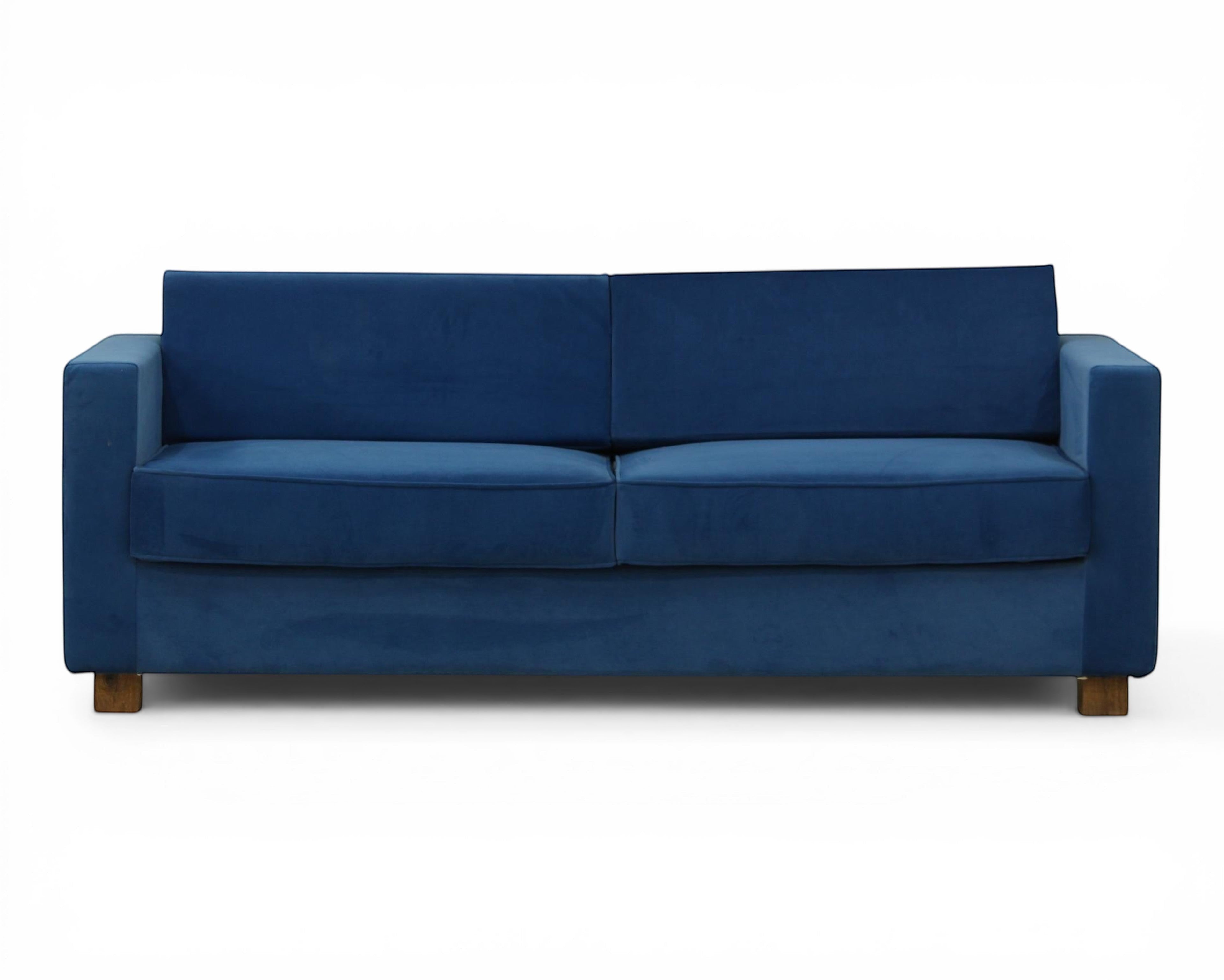 Nakoda Homes blue velvet sofa with clean lines and modern silhouette, plush upholstery, and wooden legs. Includes tan accent pillows.