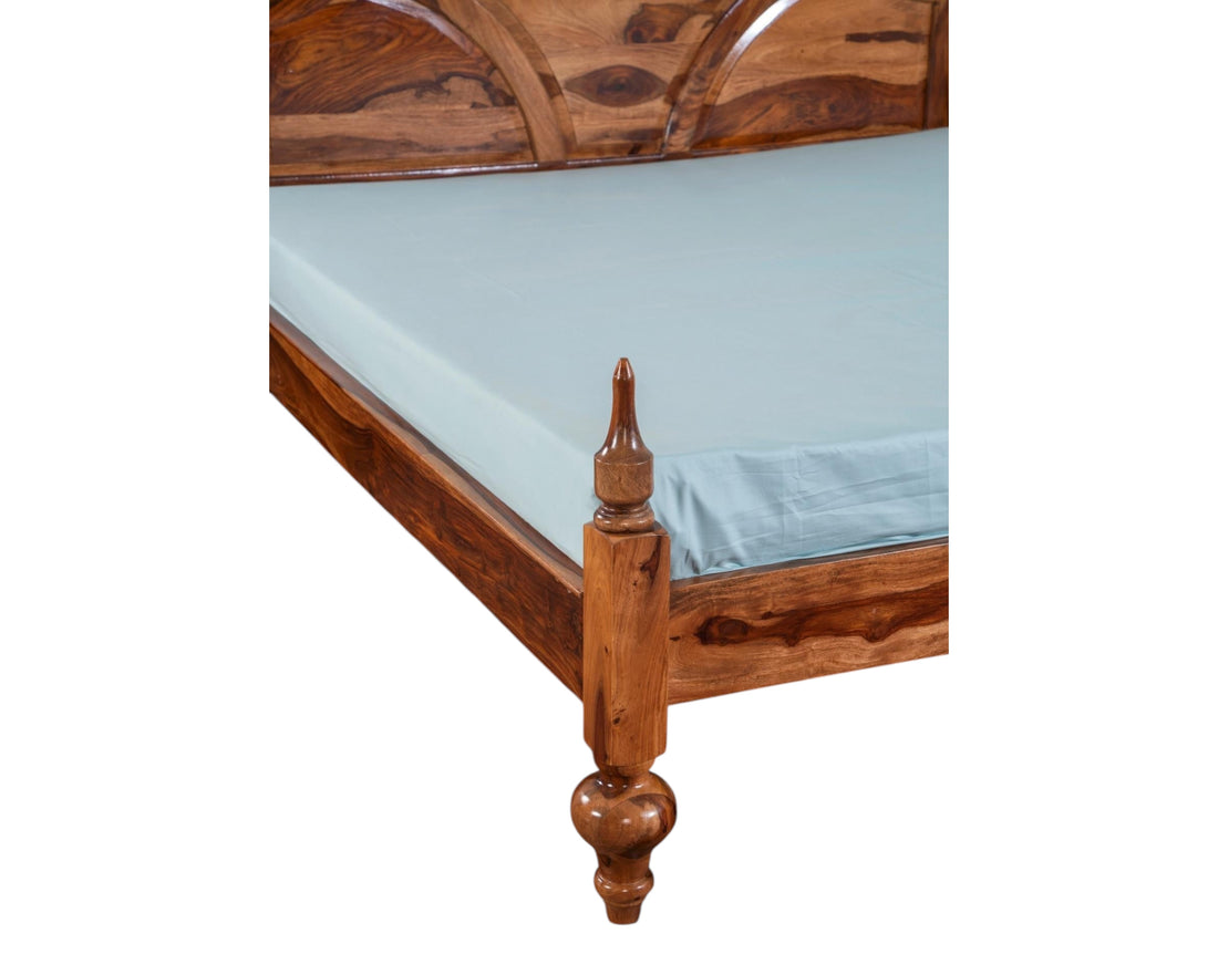 Nakoda Homes Ornate Sheesham wood bed frame with arched headboard, intricate carvings, and turned posts. Warm wood tone.